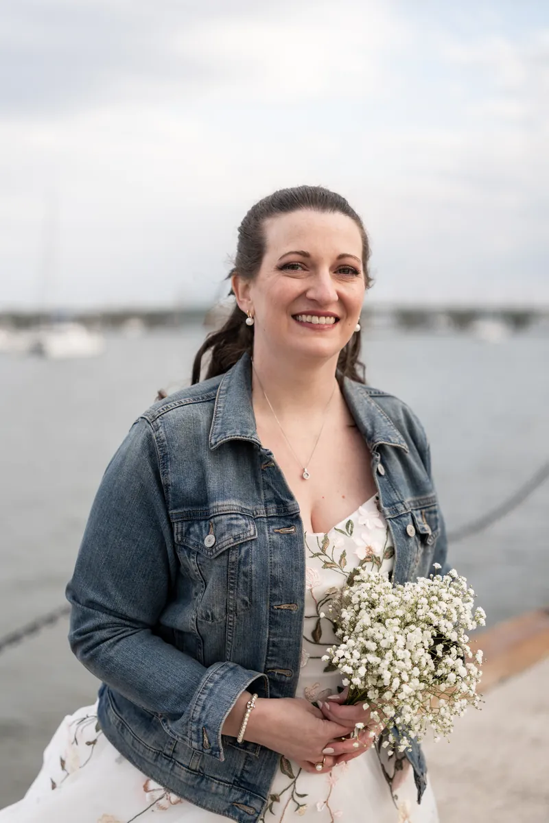 The bride in a denim jacket smiling at the camera, holding her baby's breath bouquet with the bayfront behind her