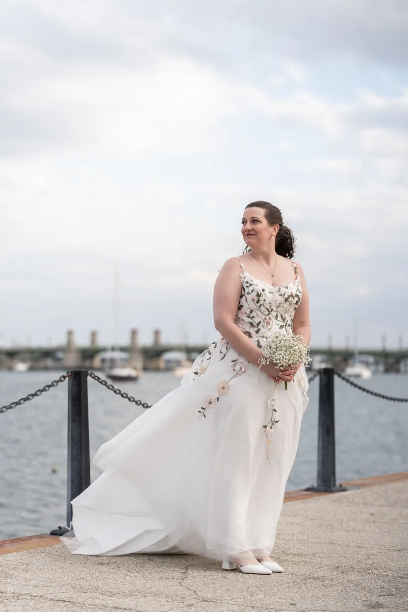 The bride standing on the bayfront dock holding her bouquet, her floral dress blowing in the breeze with the Bridge of Lions behind her