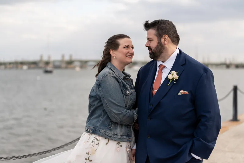 Anne and Chase looking at each other on the bayfront dock, the bay and Bridge of Lions behind them