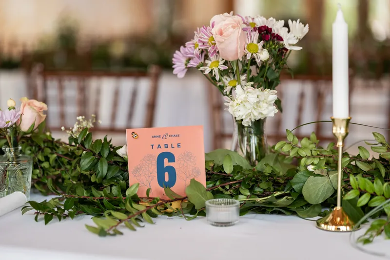 A table number card reading Anne and Chase Table 6 surrounded by greenery, flowers, and gold candlesticks