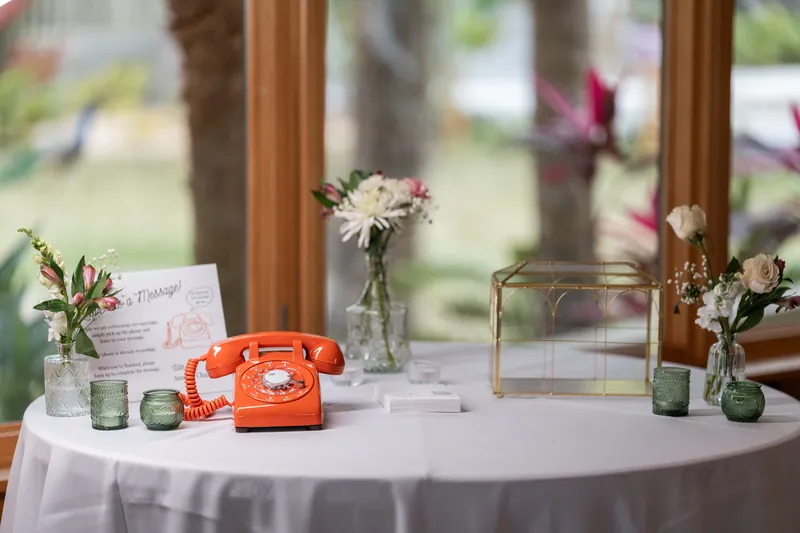 A vintage orange rotary telephone on the guest book table with bud vases, a gold card box, and a sign inviting guests to leave a message