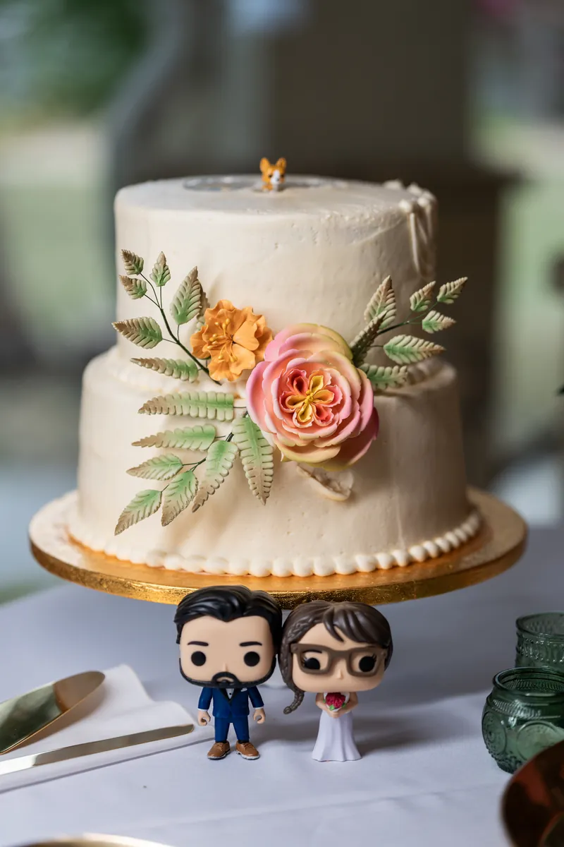 The two-tier wedding cake decorated with sugar flowers and ferns on a gold stand, with custom Funko Pop bride and groom figures in front