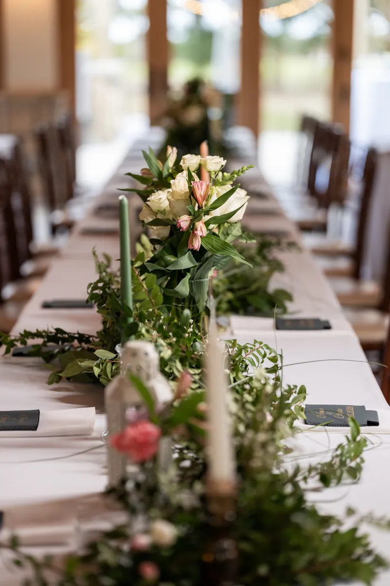 A long reception table lined with lush greenery, pink and white flowers, sage candles, and personalized place settings