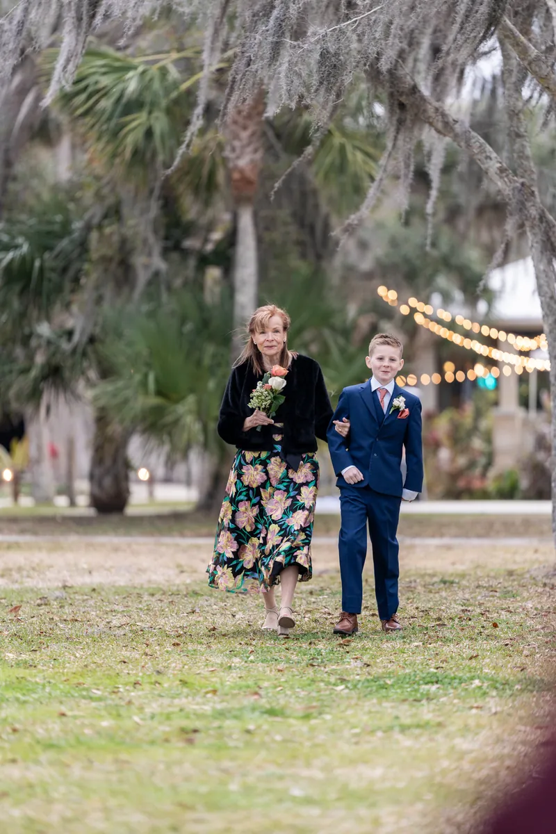 A woman in a floral skirt escorted down the aisle by a young ring bearer in a navy suit, Spanish moss overhead