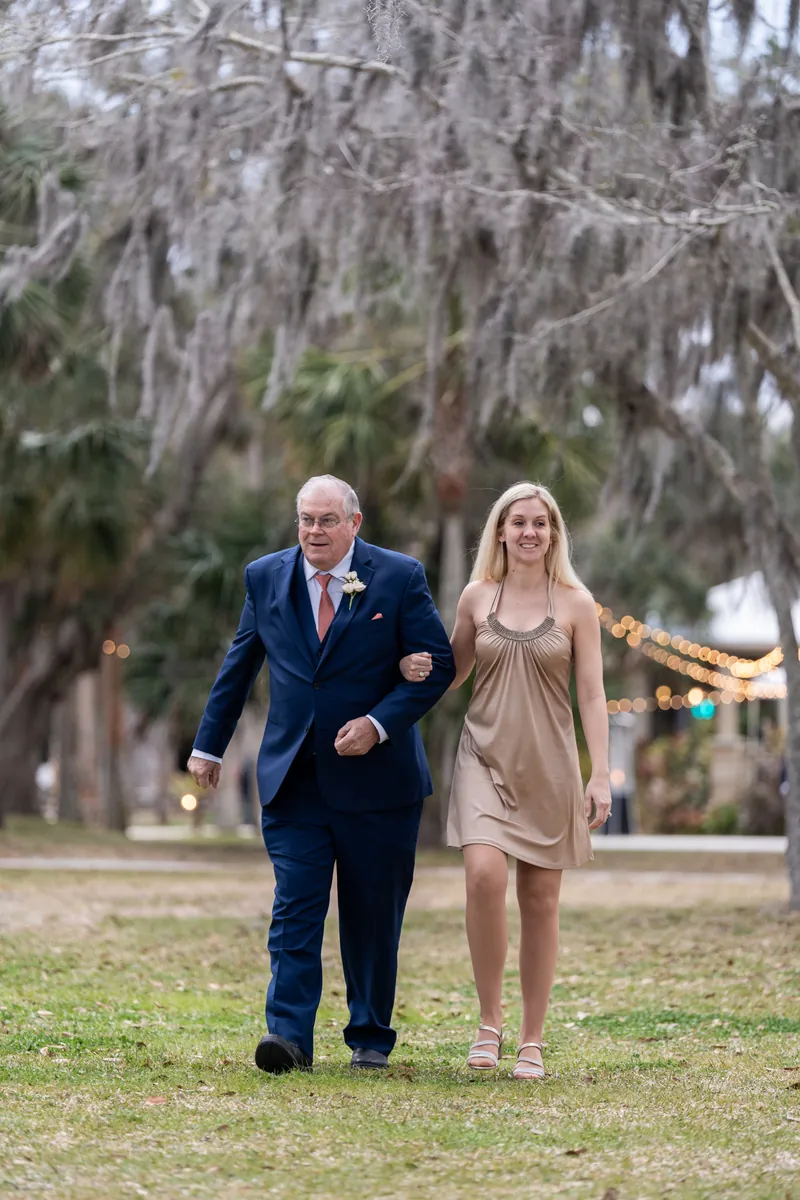 An older gentleman in a navy suit escorting a young woman in a gold dress down the ceremony aisle under moss-draped trees