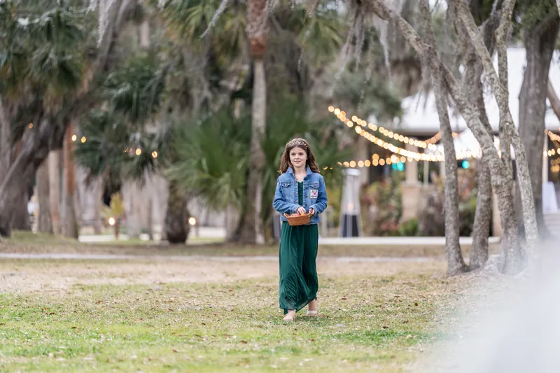 A flower girl walking down the aisle in a green dress and denim jacket, carrying a basket among palm trees and string lights