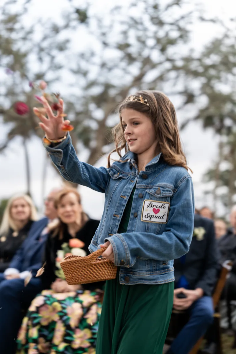 Flower girl Iris tossing petals down the aisle in her Bride Squad denim jacket as guests look on