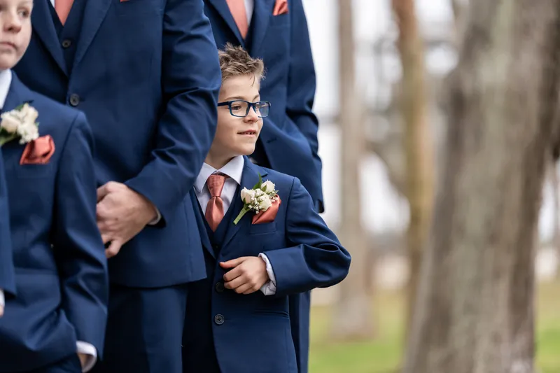 A young ring bearer in glasses standing among the groomsmen during the ceremony, matching their navy suits