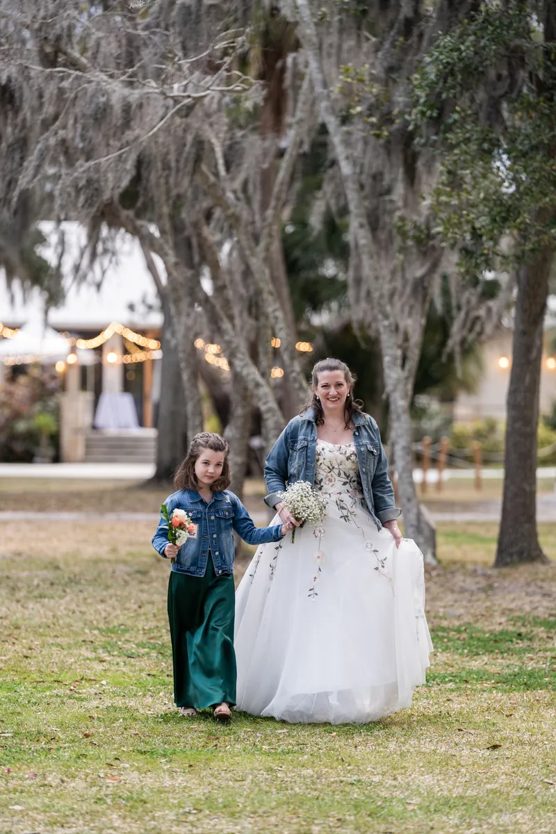 Anne and Mara walking together toward the altar, the bride in her denim jacket and floral dress with Spanish moss framing the scene