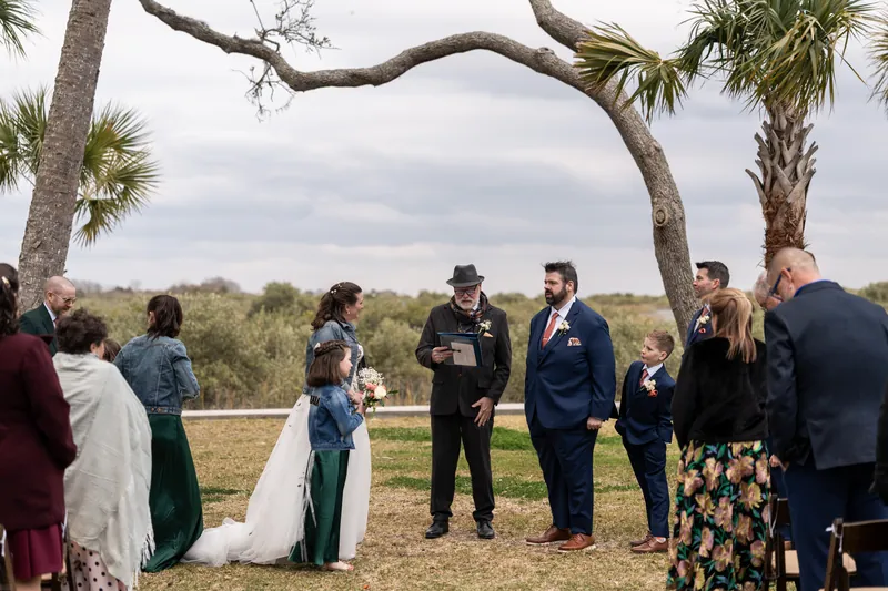 Anne holding hands with her daughter Mara at the ceremony as the wedding party and guests look on beneath palm trees