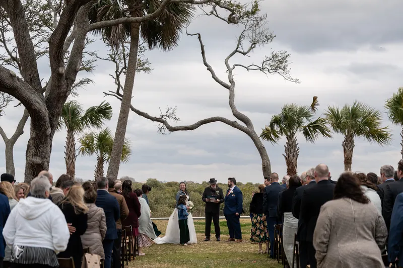 Wide view of the outdoor ceremony at Fountain of Youth with guests seated beneath twisting palm trees and a dramatic cloudy sky