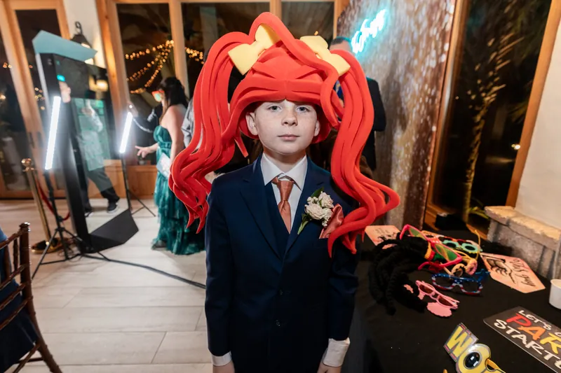 A young ring bearer hamming it up at the photo booth wearing an oversized red foam wig, props table beside him