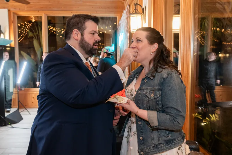 Chase feeding Anne a bite of wedding cake during the cake cutting, both laughing inside the warm reception pavilion