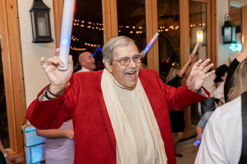 An older gentleman in a red blazer and white scarf dancing with arms spread wide, waving a glow stick during the reception