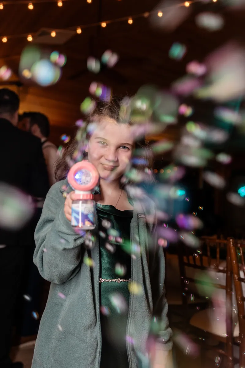 A flower girl aiming a bubble machine at the camera, colorful bubbles filling the frame with string lights behind her