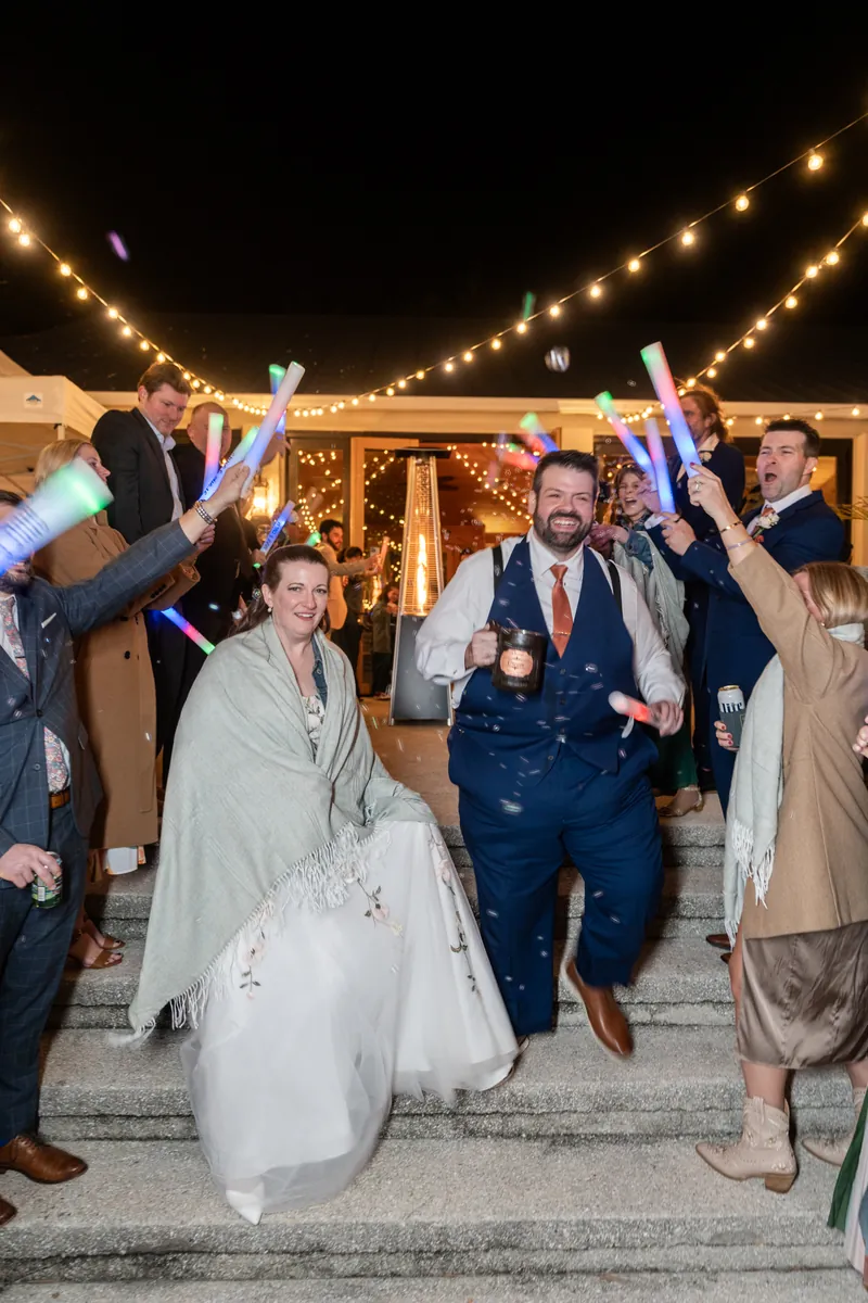 Anne and Chase making their grand exit through a tunnel of waving glow sticks, the bride wrapped in a cozy shawl and confetti falling around them