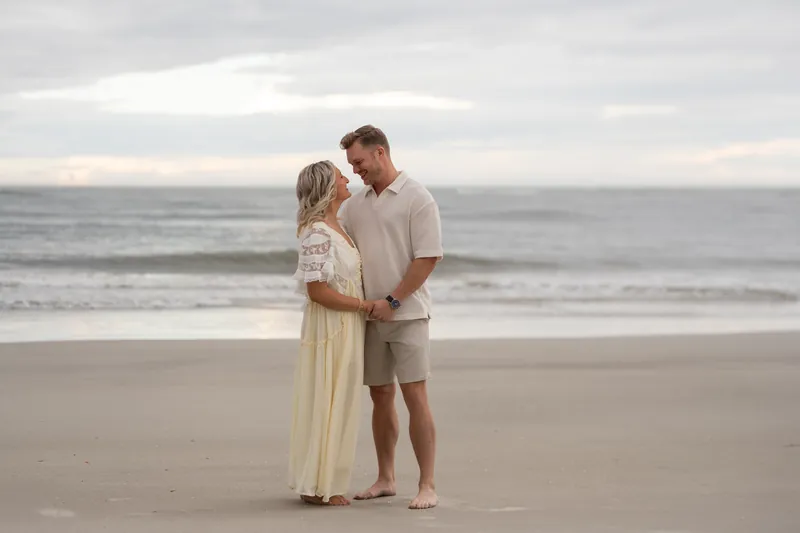 Ashley in a pale yellow dress and Craig in a tan shirt embrace on the wet sand at Vilano Beach with the ocean behind them.