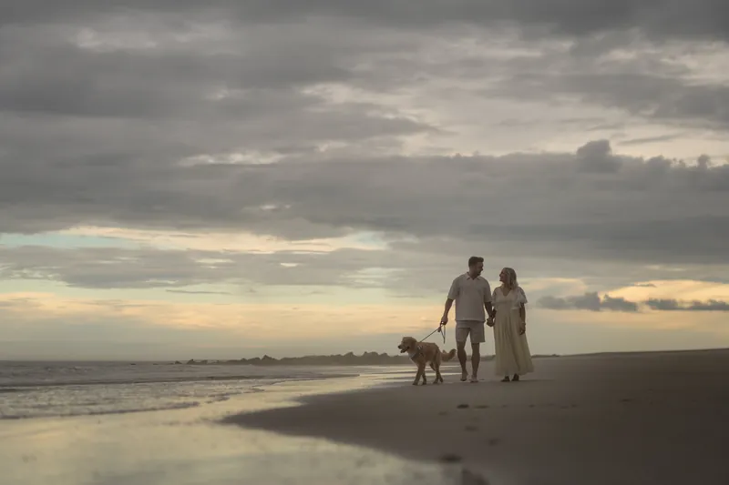 The couple and Butter walk along the shoreline at sunset, silhouetted against the clouded sky reflecting on the wet sand.