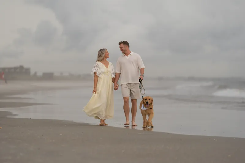 Ashley and Craig hold hands while walking along Vilano Beach with Butter, their golden-brown dog, under overcast skies.