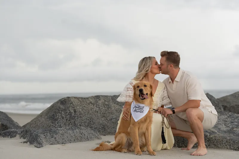 Ashley and Craig crouch beside Butter, who wears a "Fiancé" bandana, with rocky outcrops and ocean waves behind them.