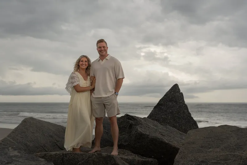 Ashley in a pale yellow dress and Craig stand together against a dramatic sky and boulders at Vilano Beach's shoreline.