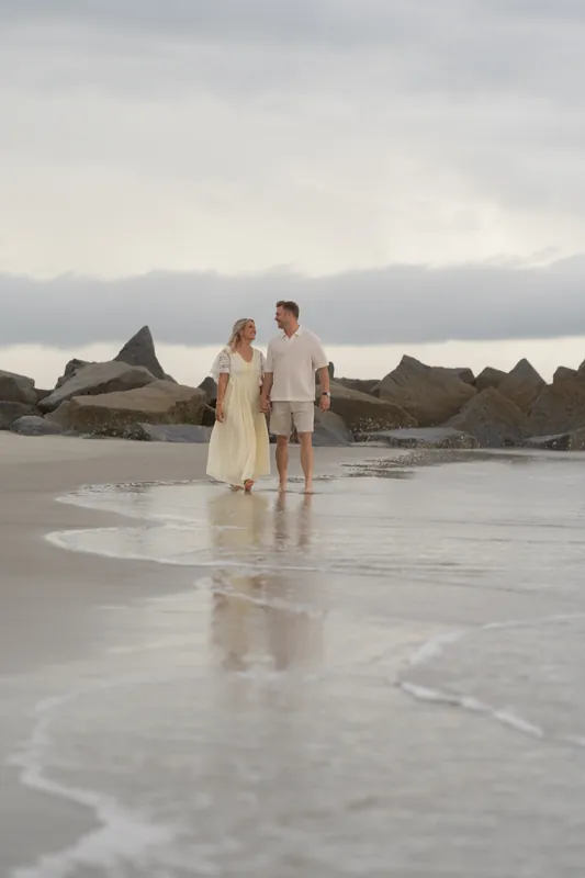 Ashley and Craig stand holding hands in shallow water at Vilano Beach with dramatic cloudy skies above and dark rock
