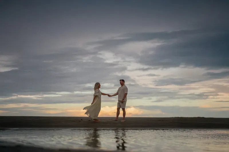 Against a colorful sunset sky, Ashley and Craig hold hands while standing in the surf at Vilano Beach.