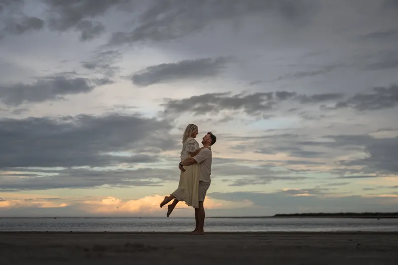 Craig lifts Ashley off her feet as they stand together on the wet sand with storm clouds stretching across the sky.