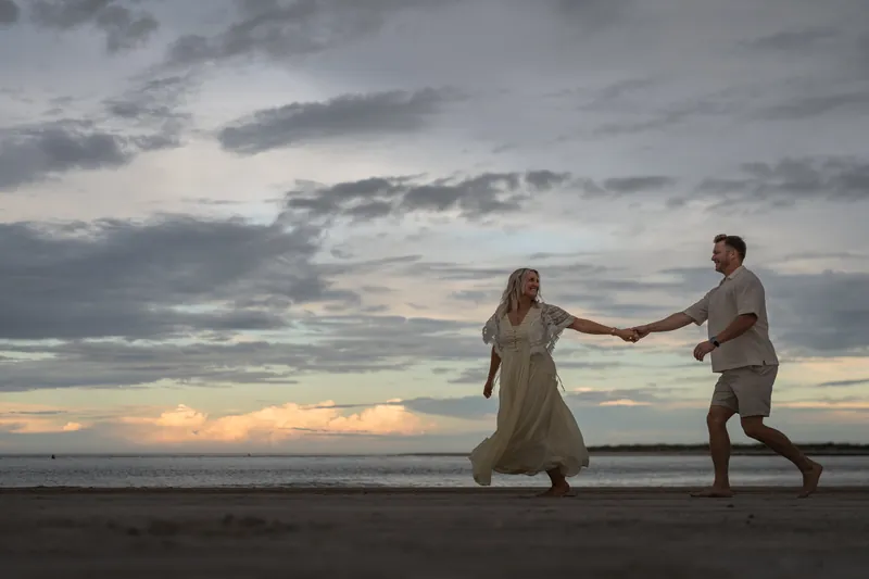 Ashley and Craig hold hands and run through shallow water at Vilano Beach under a moody, cloud-filled sunset.