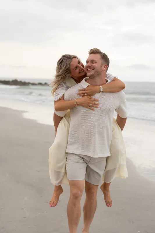 Craig carries Ashley piggyback across the sand at Vilano Beach while she wraps her arms around his shoulders.