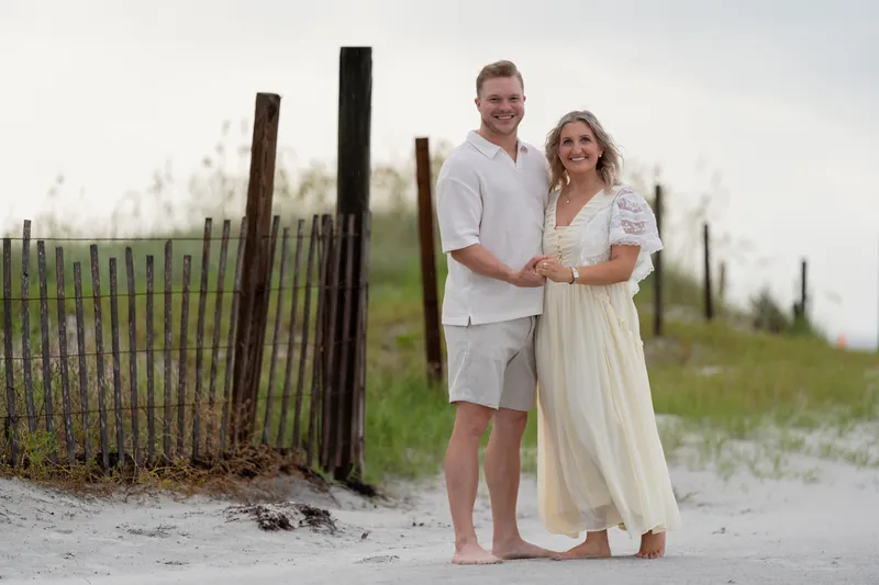 Craig and Ashley stand barefoot on the sand at Vilano Beach with a weathered fence behind them, gazing at each other