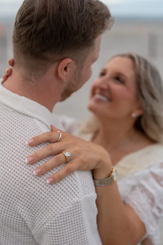 Close-up of Craig's hand on Ashley's shoulder, their engagement rings visible as they look toward each other with the beach