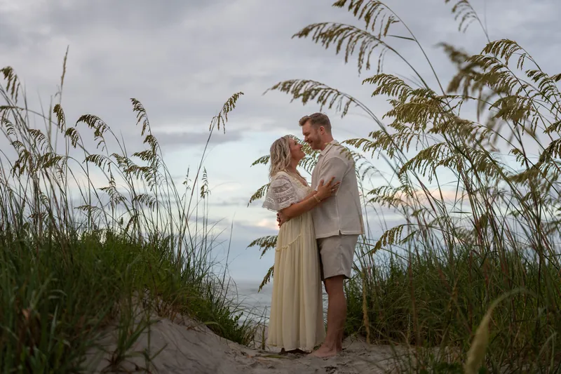 Among tall sea oats, Craig dips Ashley gently as she laughs, the Atlantic visible in the distance under cloudy skies.