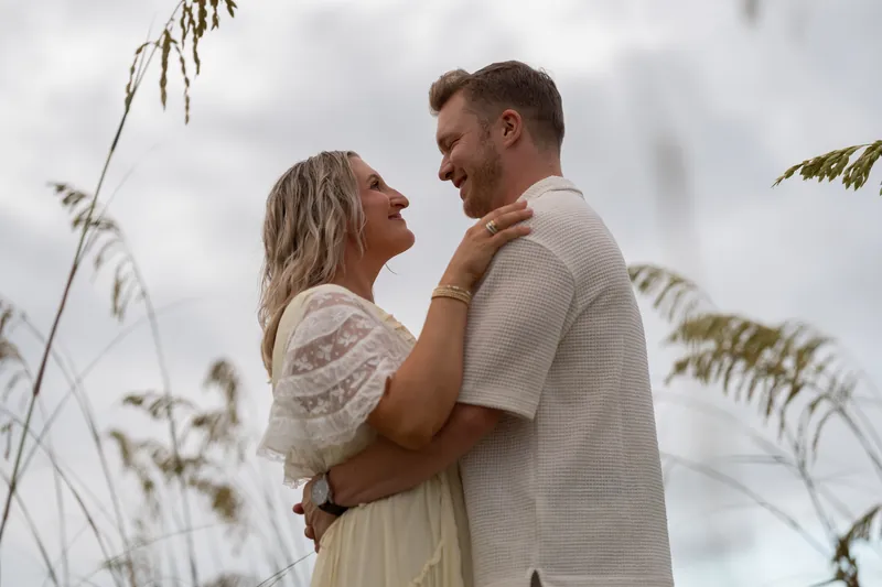 Ashley touches Craig's face while standing in beach grass, both smiling softly as wind moves the dune vegetation around them.