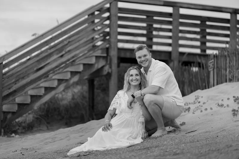 In black and white, Ashley sits on the sand leaning against Craig's leg while he looks down at her, a wooden beach access