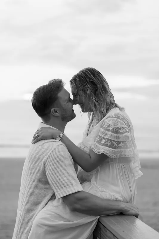 Ashley and Craig share a tender kiss on the shore, the overcast sky and ocean stretching behind them.