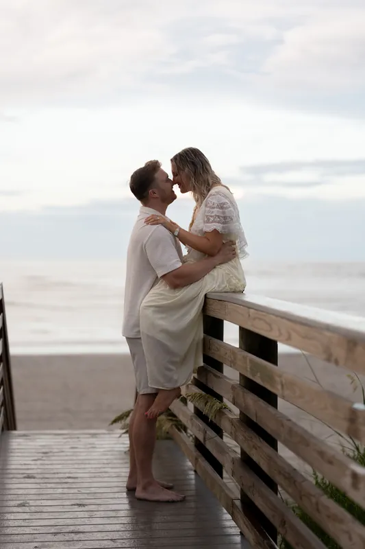 Standing on a wooden boardwalk at Vilano Beach, Craig lifts Ashley as they kiss with the beach visible below.