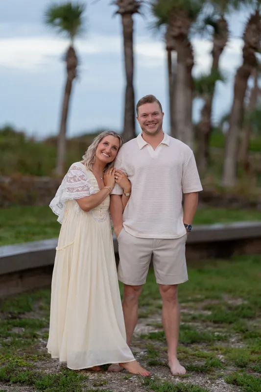 Ashley leans against Craig as they stand together among palmetto palms at Vilano Beach, both wearing light-colored clothing.