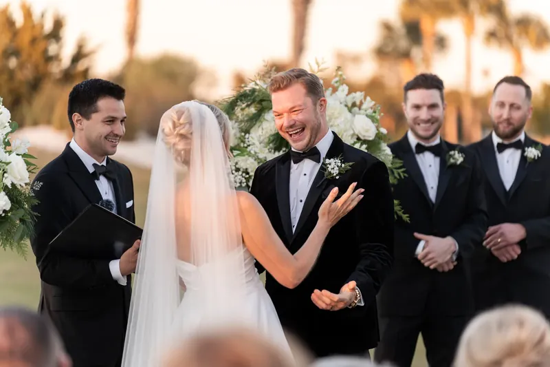 Craig laughs beside Ashley, who wears a white gown and veil, with groomsmen standing nearby.