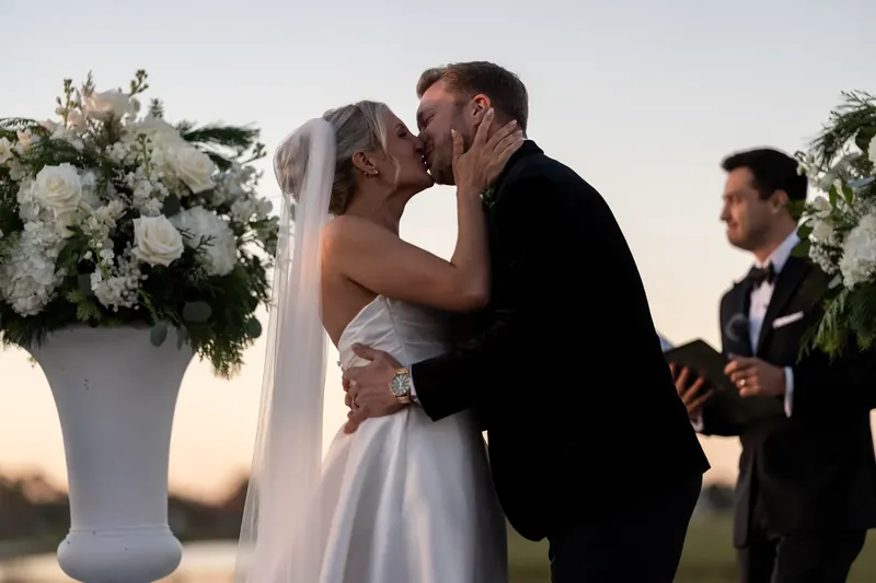 Ashley and Craig kiss at the altar with white floral arrangements on either side.