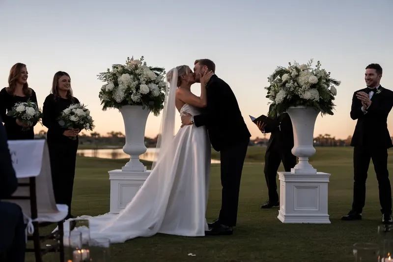 Ashley and Craig exchange vows at sunset on the golf course, surrounded by white floral arrangements and their wedding party.