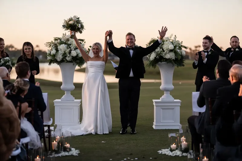 Craig raises his arms in celebration with Ashley beside him on the grass, guests seated on both sides.