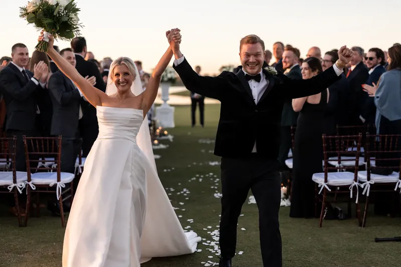 Ashley and Craig raise their joined hands in triumph as guests celebrate around them on the golf course at dusk.