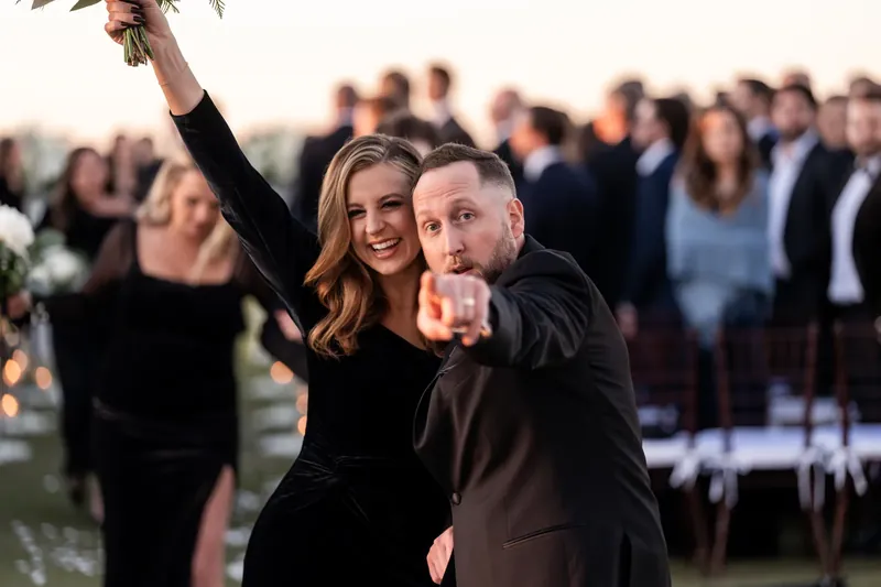 Ashley holds up her bouquet while Craig points toward the crowd during their joyful recessional as a couple.