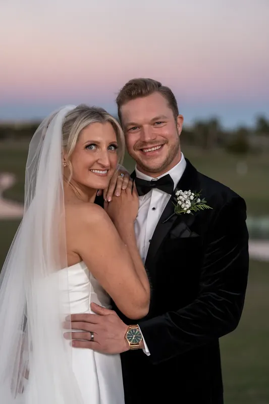 Ashley and Craig smile at dusk in their wedding attire, golf course stretching behind them.