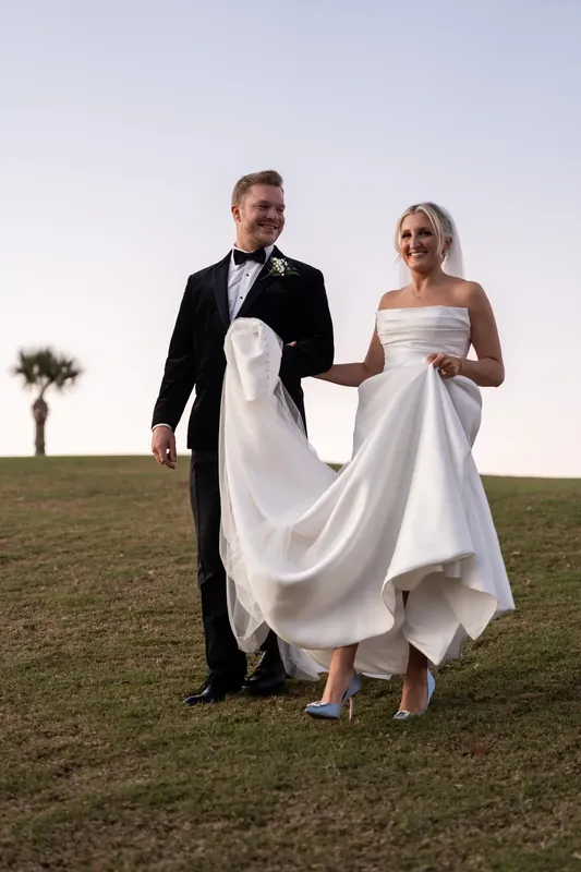 Craig and Ashley walk hand in hand across the grass, her dress trailing behind them.