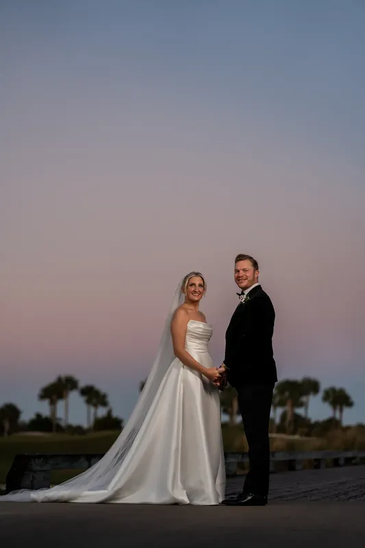 The couple stands on a wooden deck at dusk with fairways and water visible beyond.
