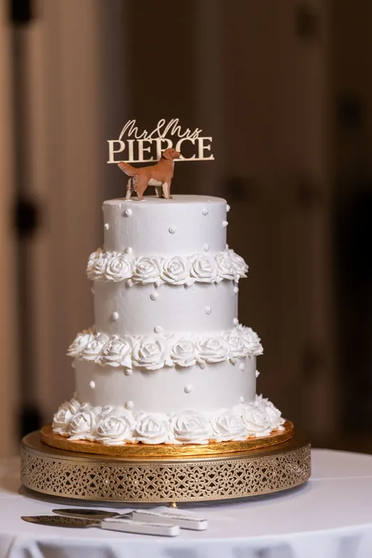 Three-tier white wedding cake adorned with rosette piping and gold-trimmed base displays an "Ashley & Craig" topper.