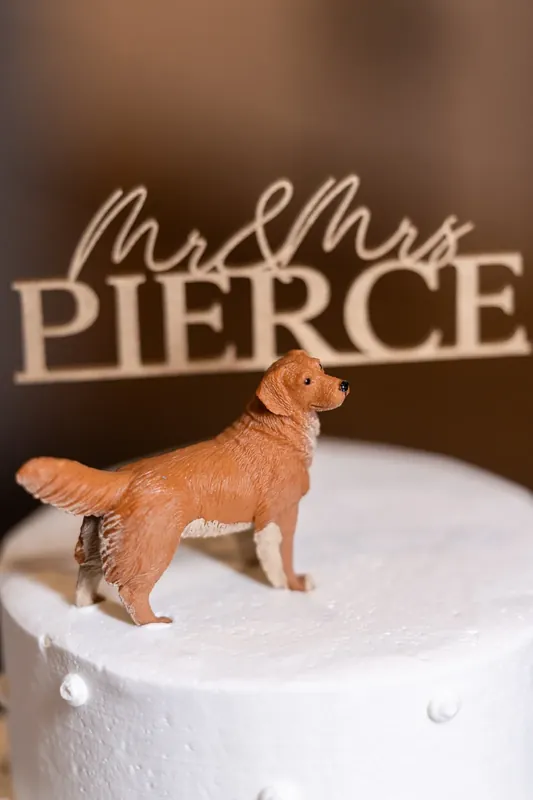 A golden dog figurine stands atop a white cake tier beside a wooden "Mr & Mrs Pierce" sign at their reception.