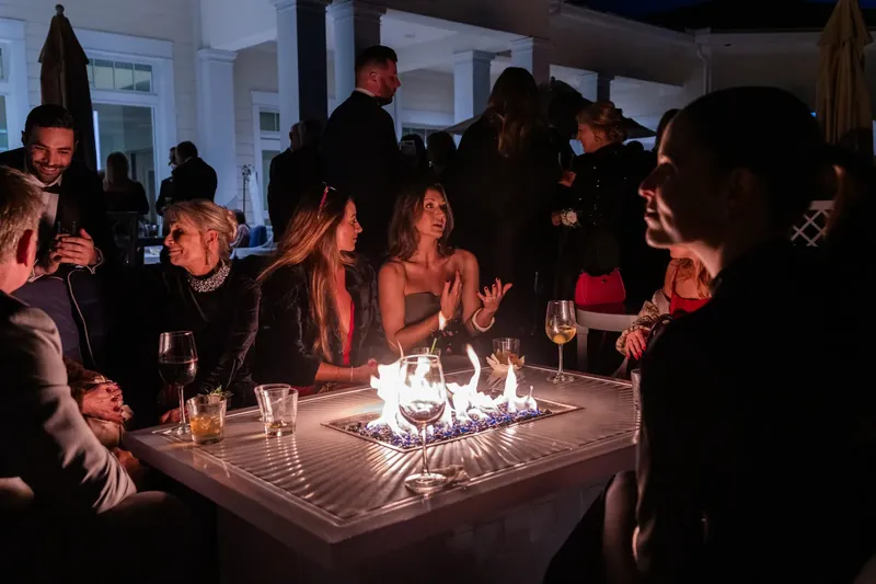 Wedding guests gather around a low fire table at night, holding drinks and conversing in warm ambient light.